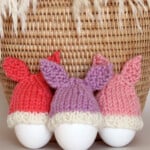 Knitted Bunny Egg Cozies in peach, purple, and pink yarn colors beside a wicker basket.