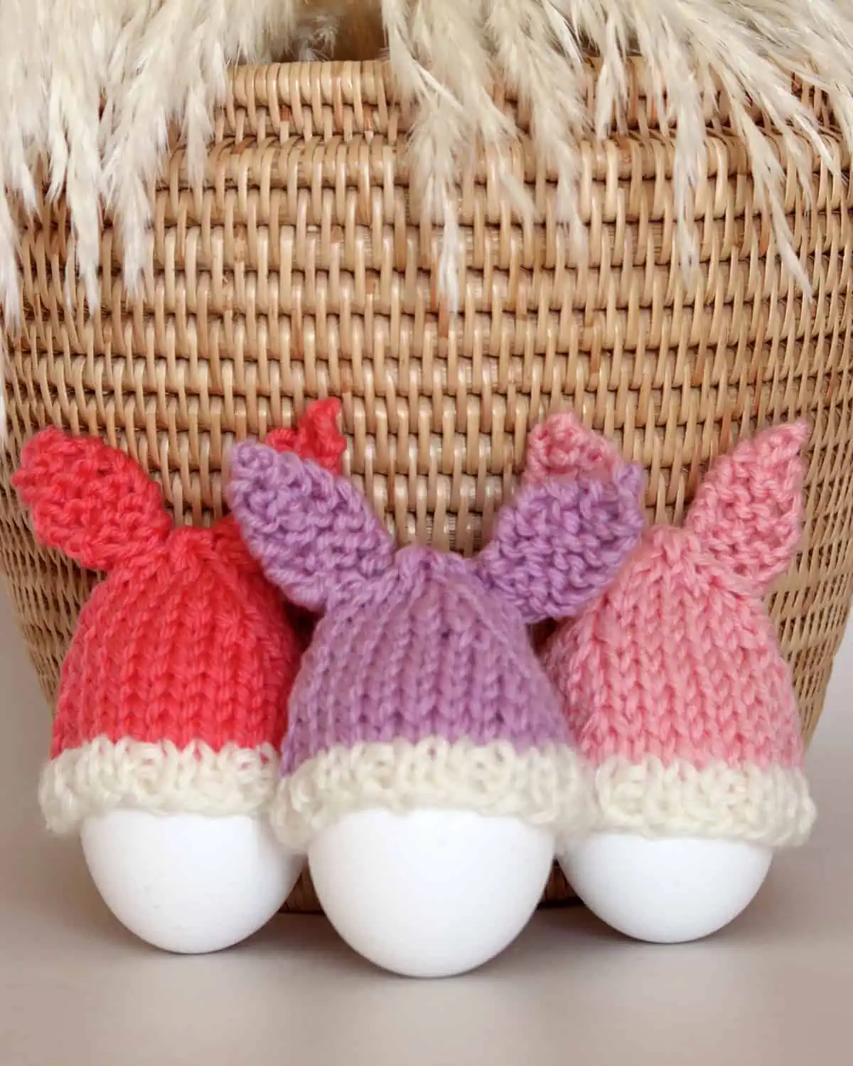 Knitted Bunny Egg Cozies in peach, purple, and pink yarn colors beside a wicker basket.