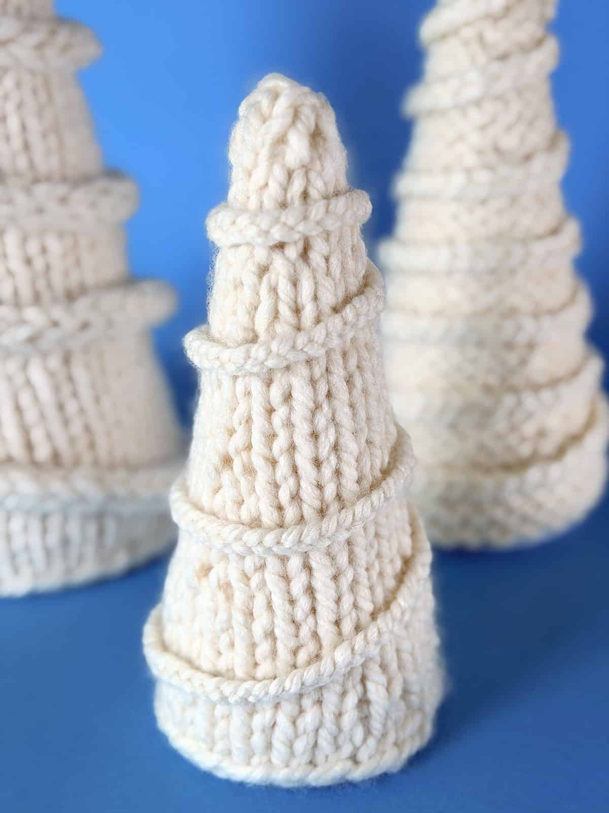 Close-up of a hand-knitted ivory Christmas tree made with thick yarn in stockinette stitch, wrapped with a spiral I-cord garland, displayed on a soft blue background.