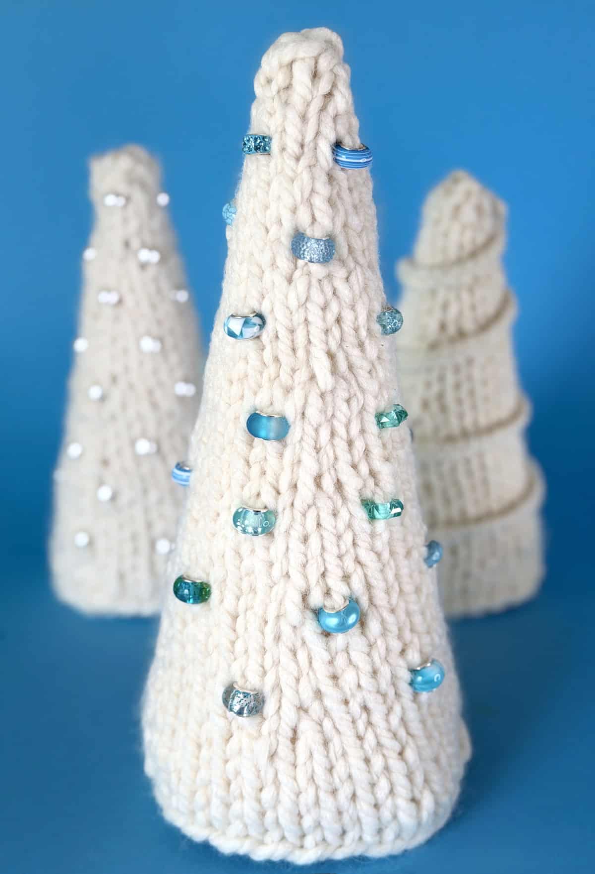 Hand-knitted ivory Christmas tree decorated with blue glass beads, displayed with two additional knit trees on a soft blue background.