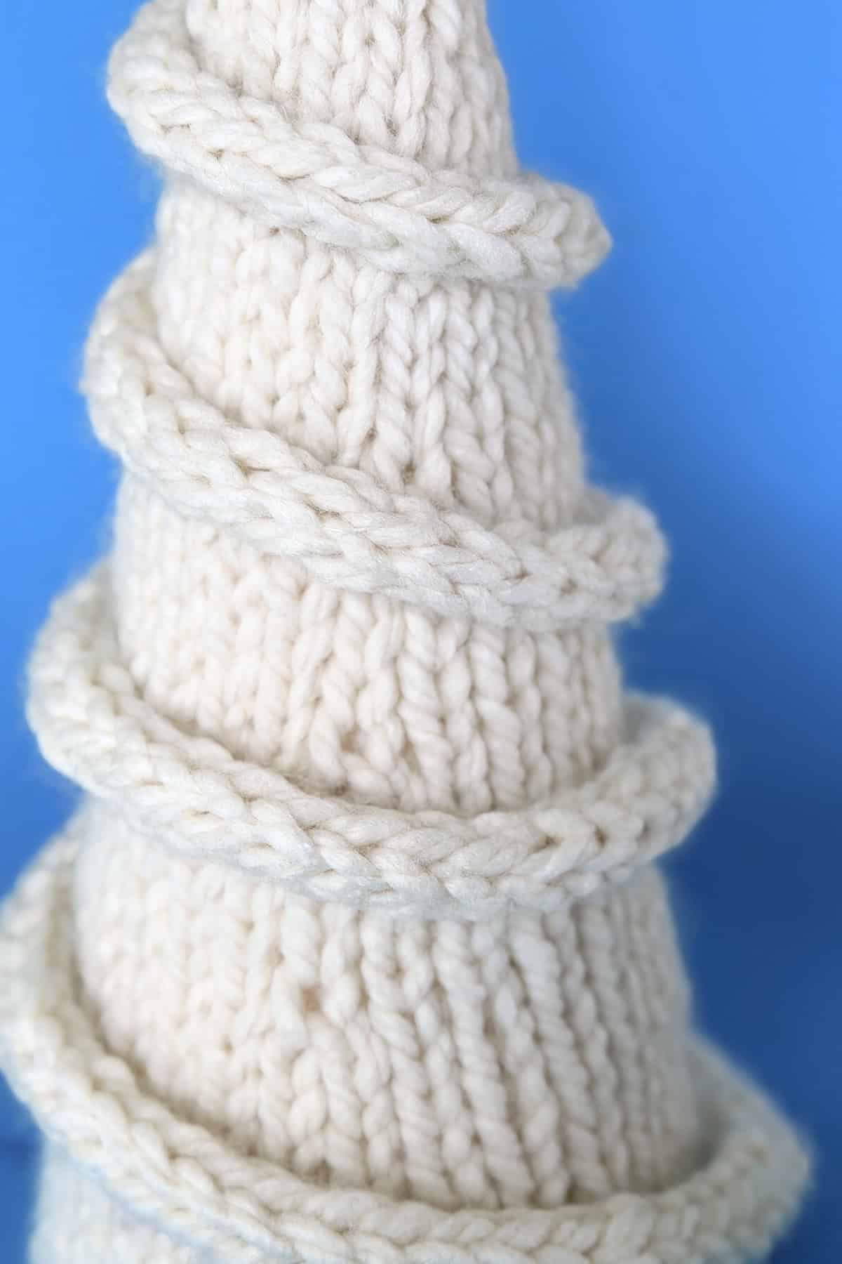 Close-up of a hand-knitted ivory Christmas tree in stockinette stitch, wrapped with a spiral I-cord garland against a soft blue background.