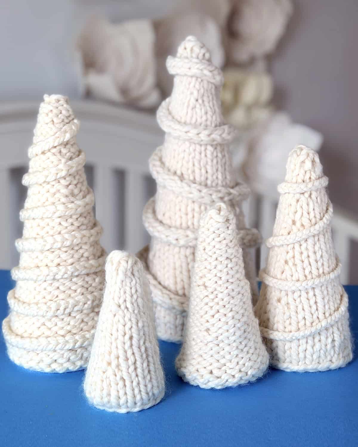 A collection of hand-knitted ivory tabletop Christmas trees displayed on a blue surface, featuring both stockinette and reverse stockinette textures with spiral I-cord garlands.