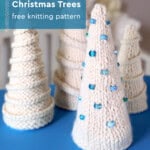A collection of hand-knitted ivory tabletop Christmas trees displayed on a soft blue surface. Each tree features a unique texture, from smooth stockinette to ridged purl patterns, with some decorated using spiral garlands and blue glass beads.