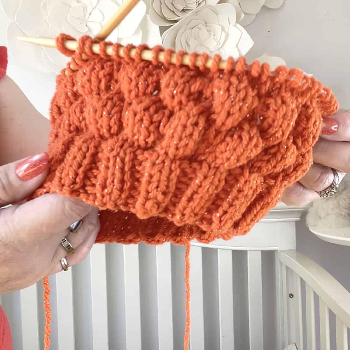 Hands holding a Bubble Beanie hat in progress on circular needles, knitted with orange sparkly yarn in a raised textured stitch.