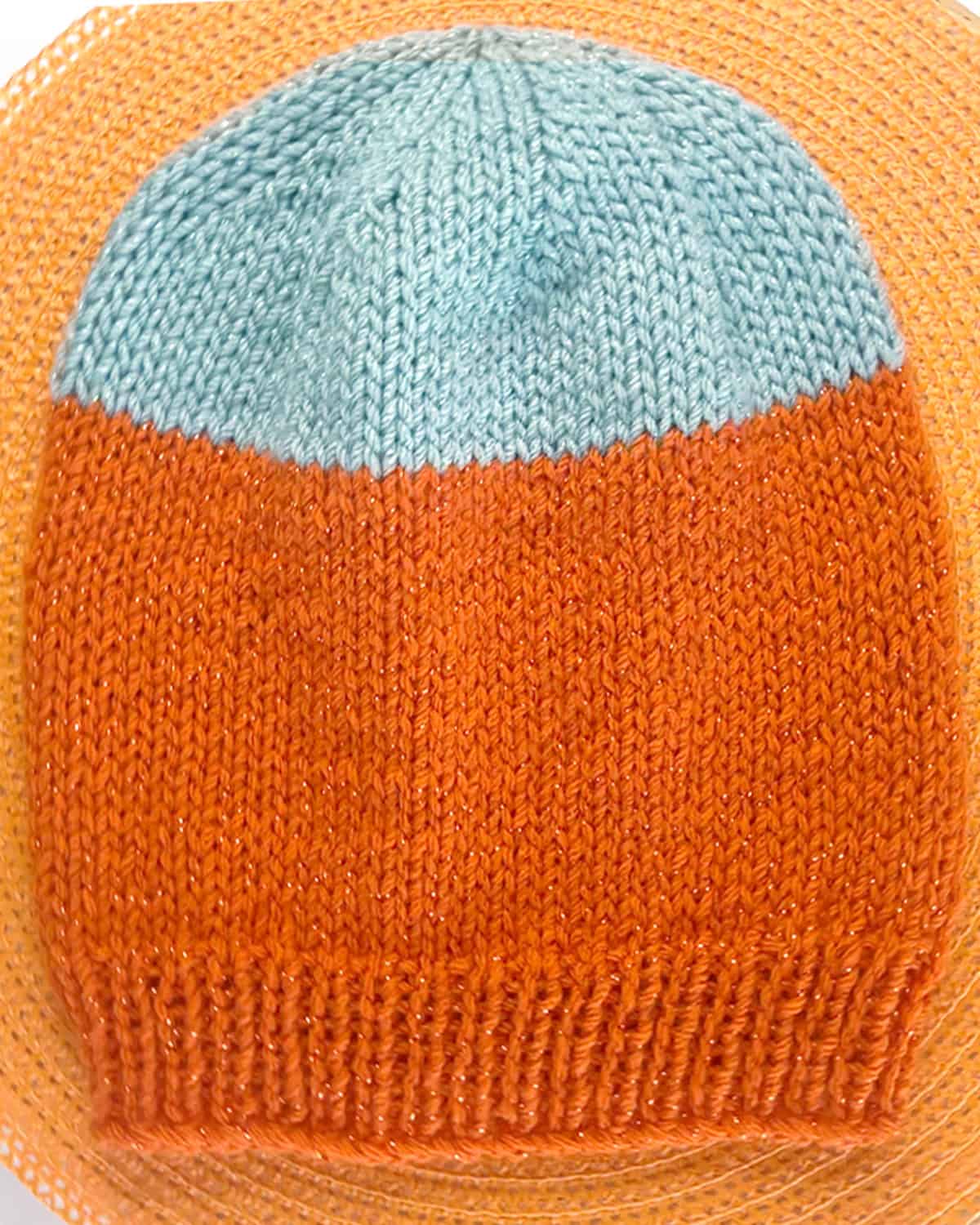 Hand-knit two-tone slouchy beanie hat in orange sparkly yarn with a light blue crown, shown flat on a woven mat.