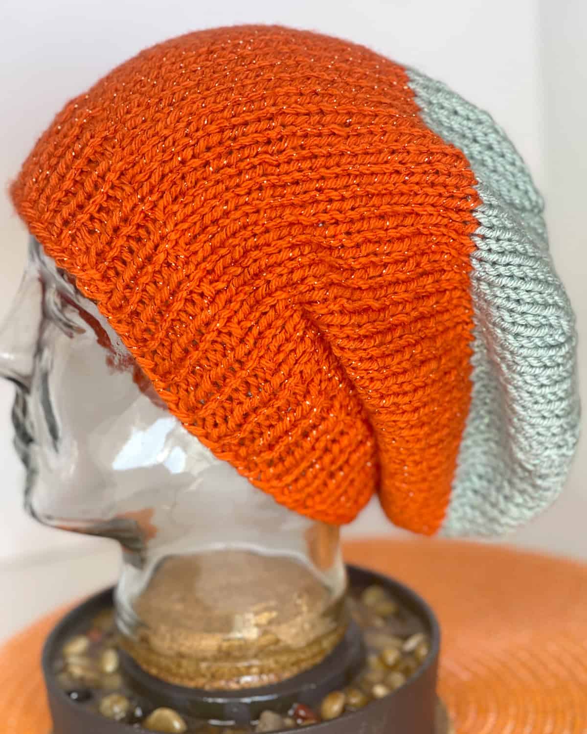 Knitted slouchy beanie hat in sparkly orange yarn with gray accent, displayed on a glass mannequin head.