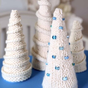 A collection of hand-knitted ivory tabletop Christmas trees displayed on a soft blue surface. Each tree features a unique texture, from smooth stockinette to ridged purl patterns, with some decorated using spiral garlands and blue glass beads.