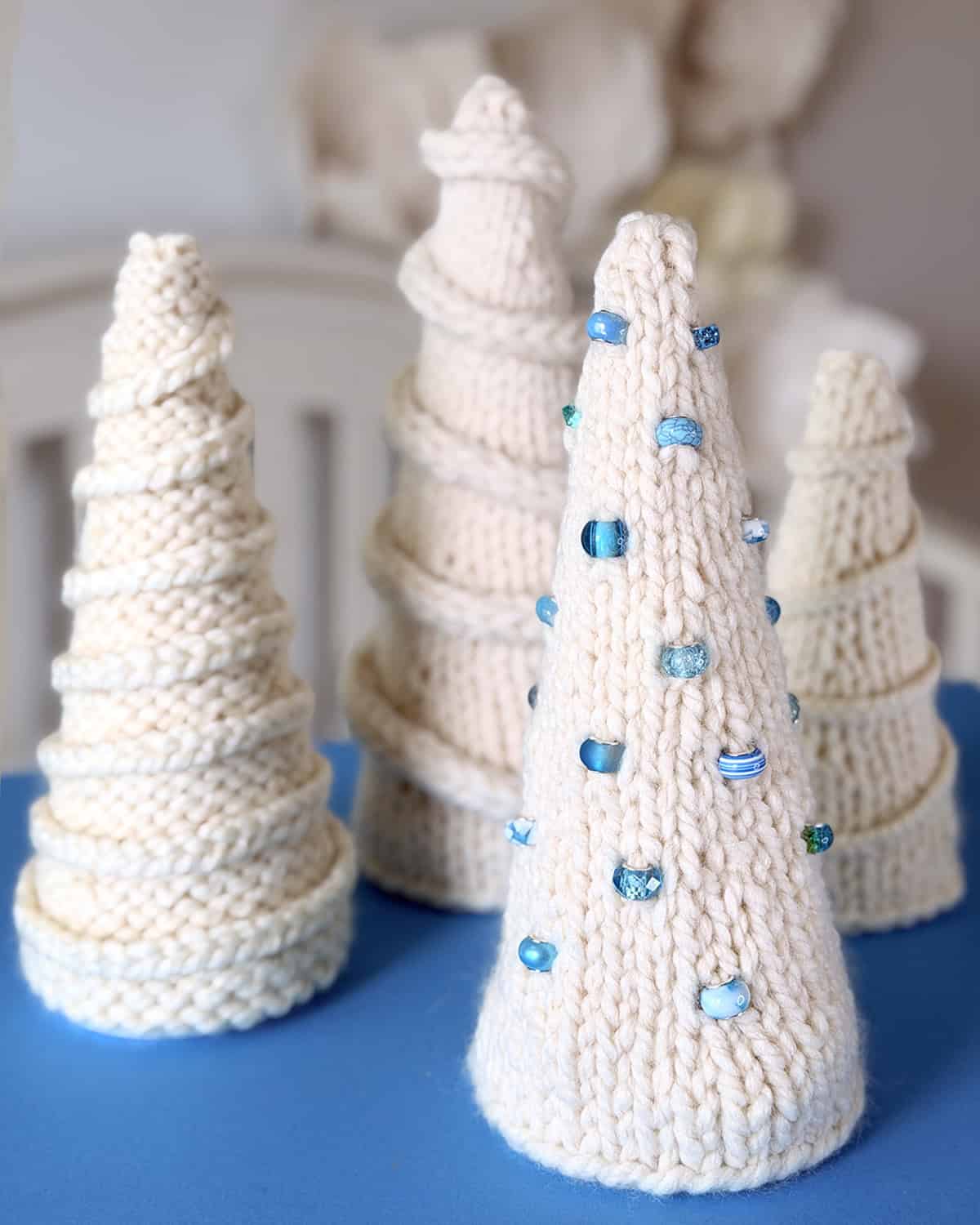 A collection of hand-knitted ivory tabletop Christmas trees displayed on a soft blue surface. Each tree features a unique texture, from smooth stockinette to ridged purl patterns, with some decorated using spiral garlands and blue glass beads.