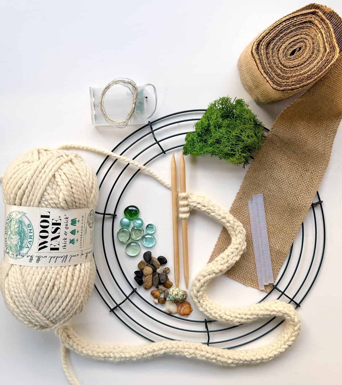 Craft supplies for Tree of Life wreath including chunky yarn, wreath form, knitting needles, moss, stones, burlap ribbon, and craft lights.