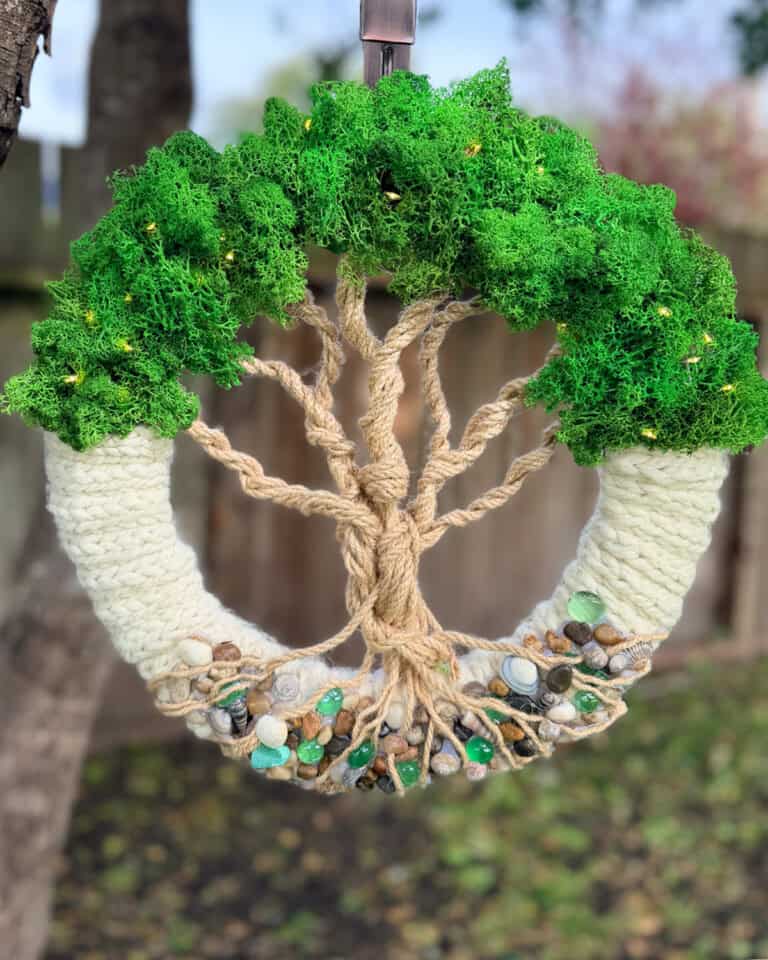 Knitted Tree of Life wreath hanging outdoors with moss greenery, rope branches, and stone base.