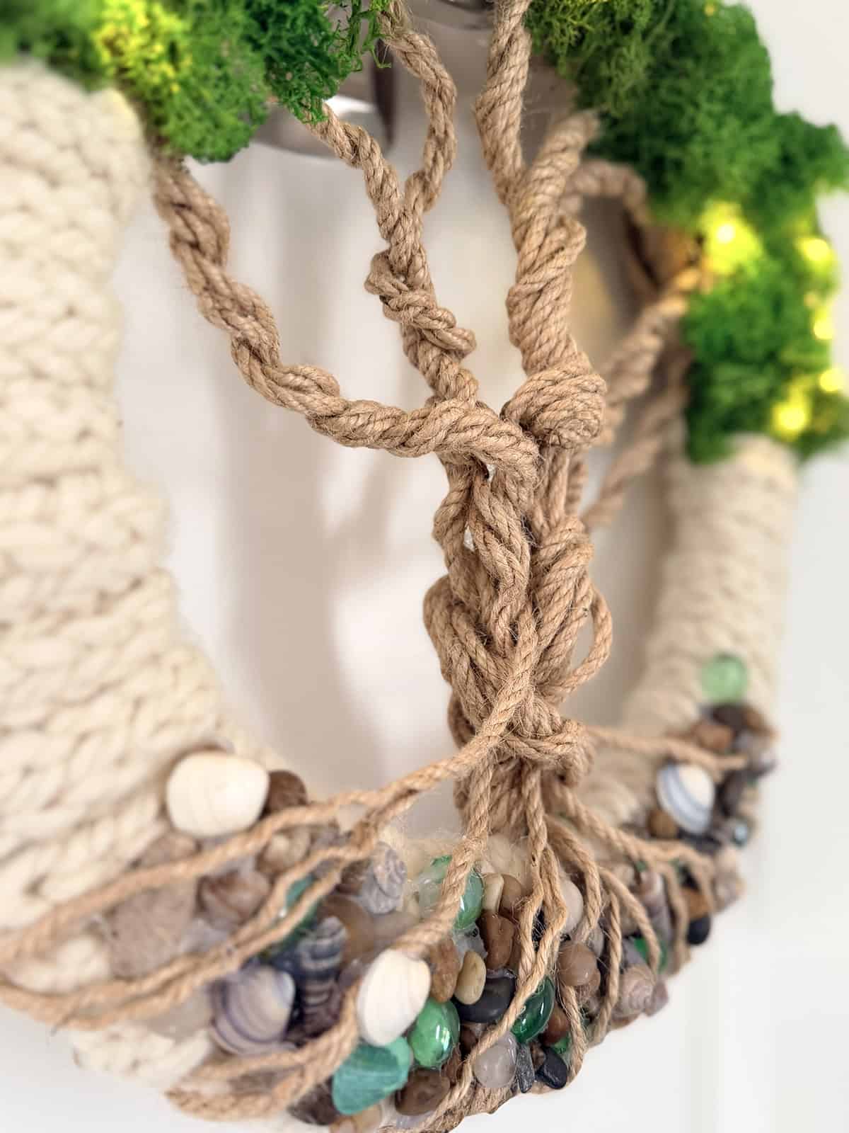 Close-up of Tree of Life wreath showing twisted jute rope trunk and roots over stones, shells, and green glass gems with knitted base and moss foliage above.