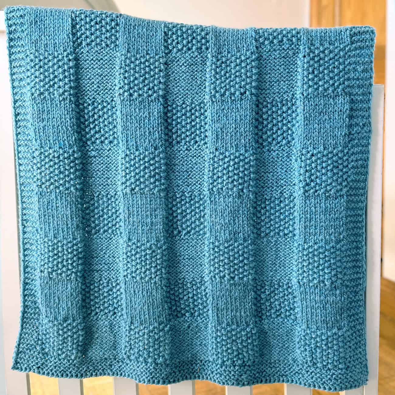 Blue Pescadero Harvest Blanket draped over a crib, showing the reversible checkerboard knit texture.